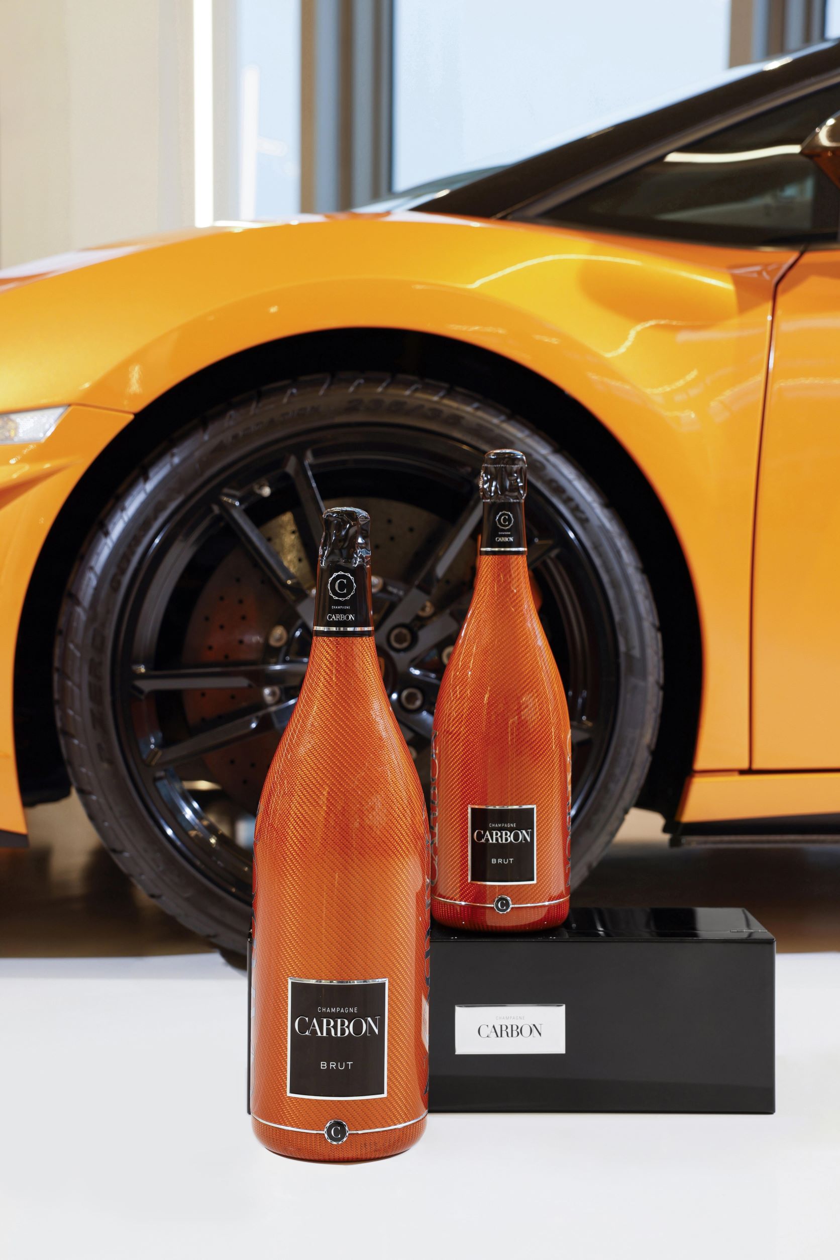 Two orange bottles of Champagne Carbon presented in front of a similar coloured Lamborghini supercar