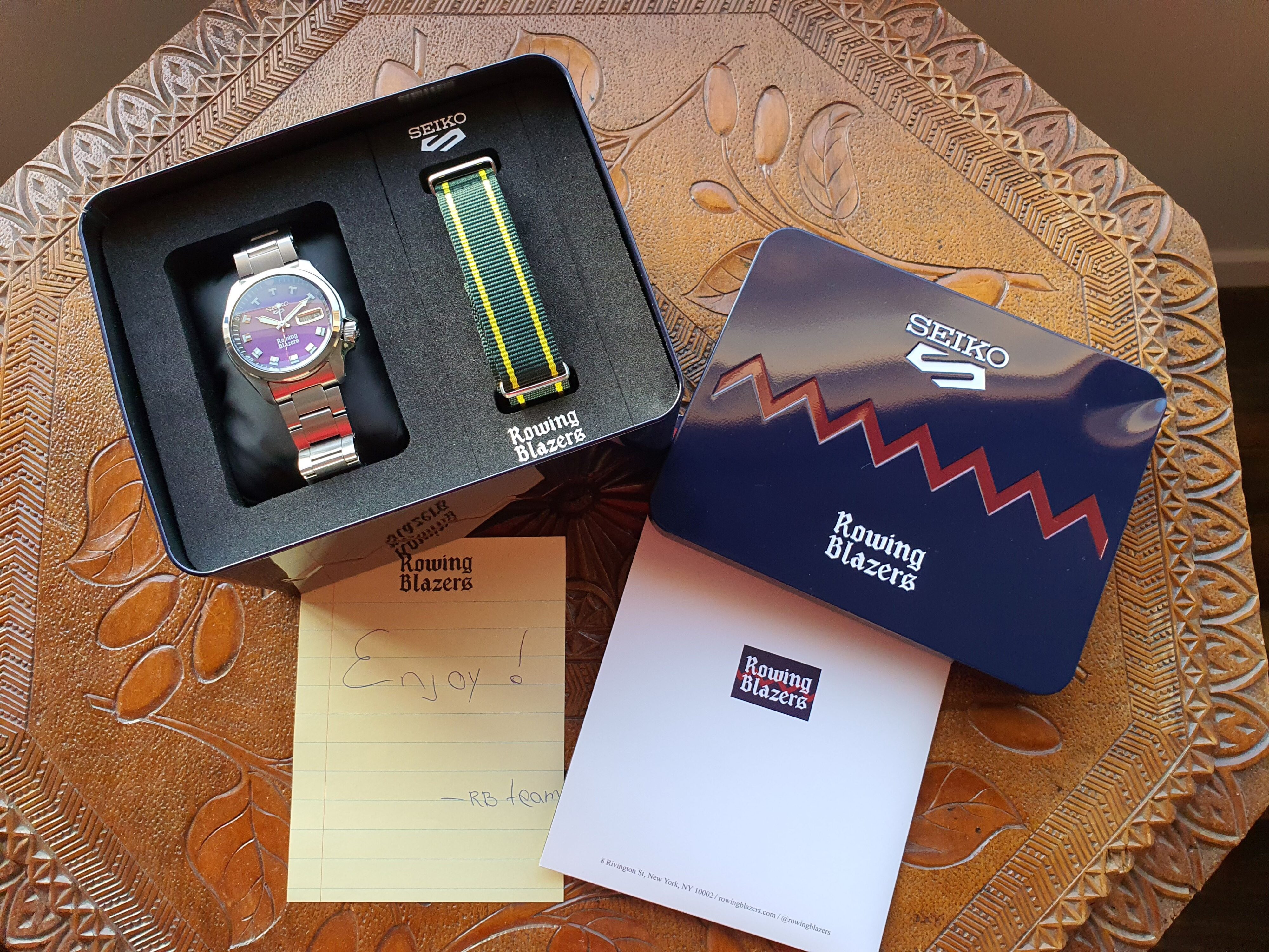 A photo of an open metal display box featuring the Seiko 5 x Rowing Blazers purple dial wristwatch, NATO strap, handwritten note and document folder
