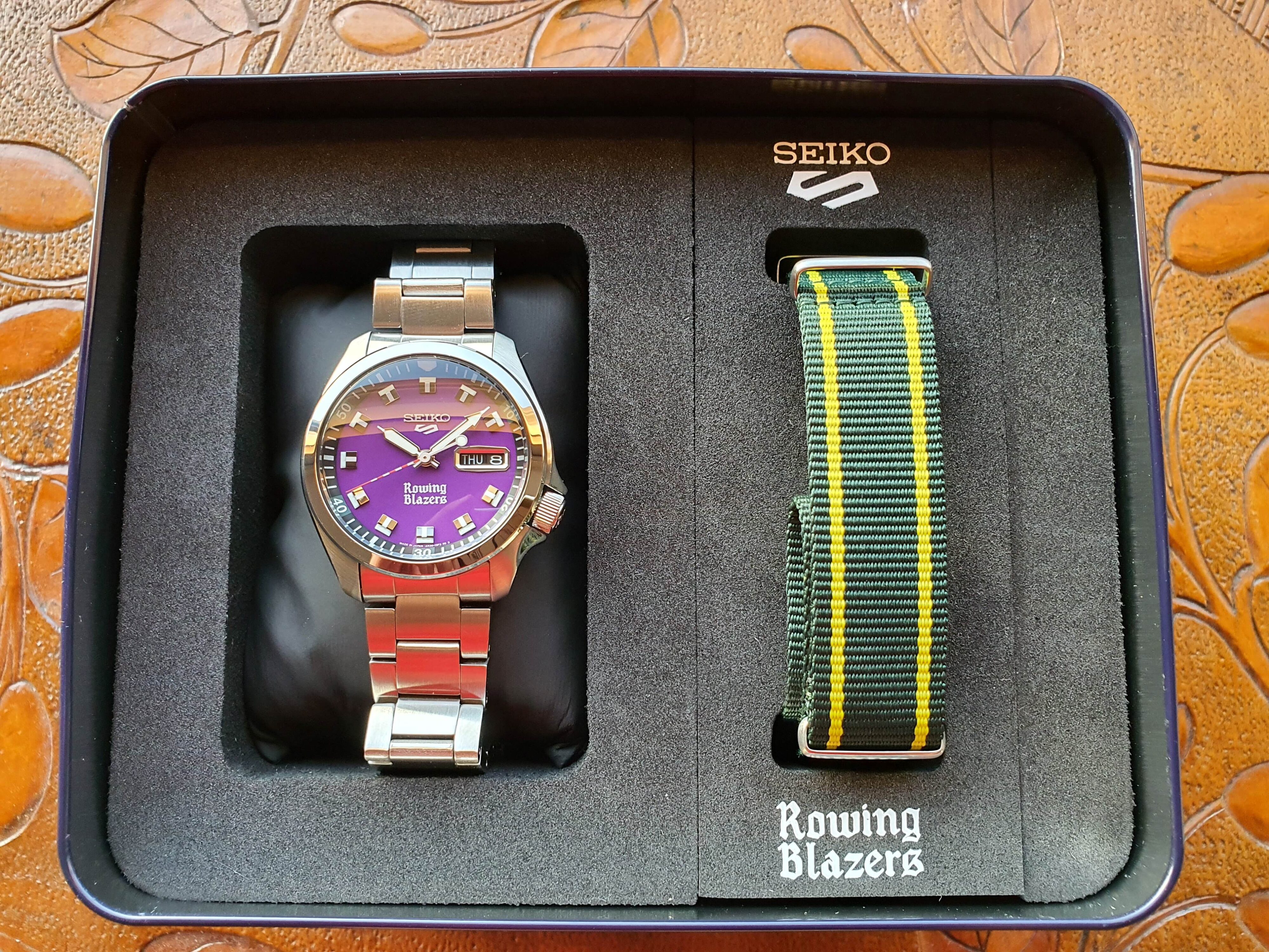 Photo of an open Seiko 5 x Rowing Blazers watch display case showing a purple dial version of the collaboration wristwatch and the accompanying nato strap in green and yellow.