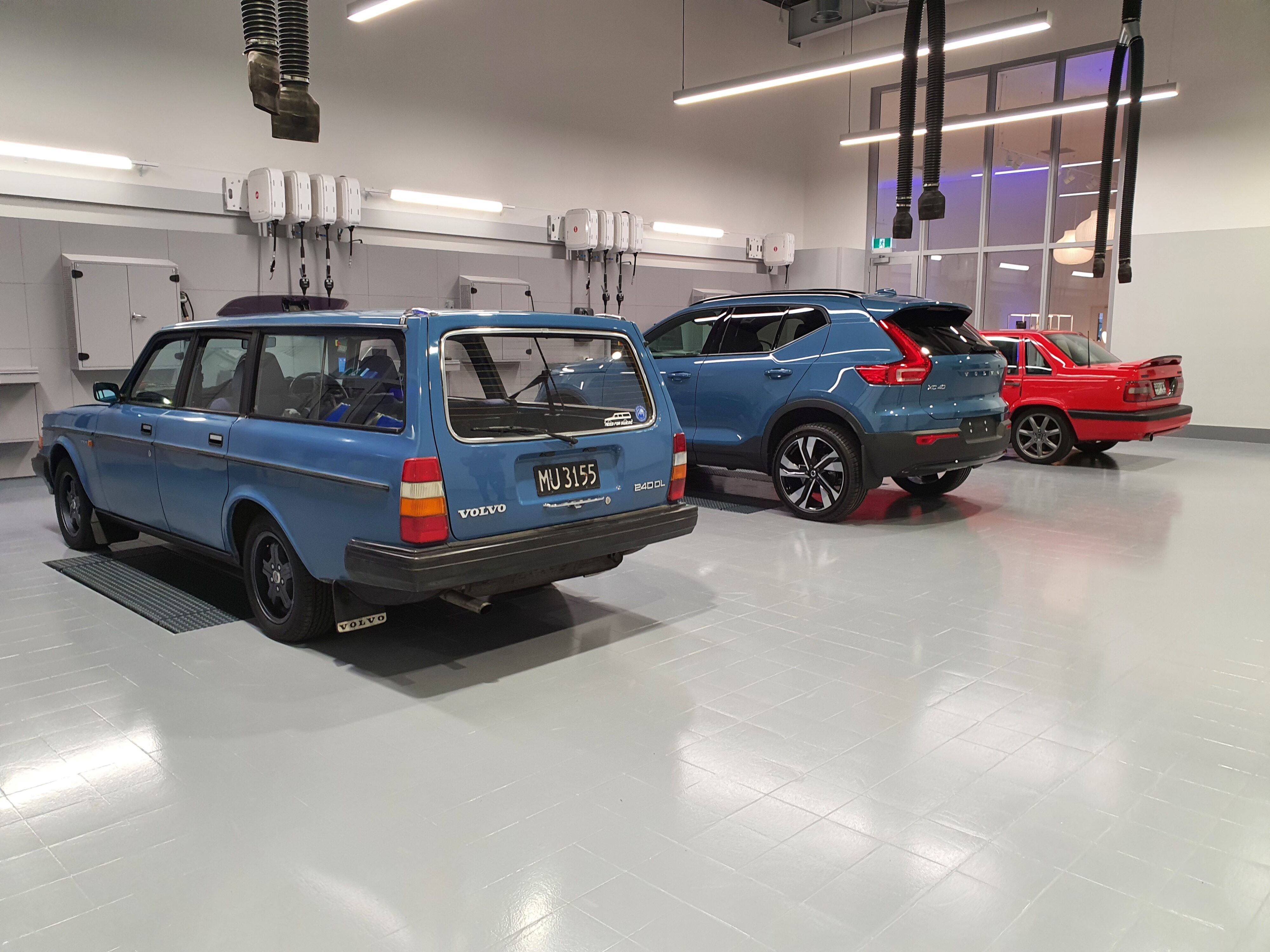 Three Volvo cars present in the workshop of the showroom. From left, a 246GL in Fjord Blue, a XC40 in Fjord Blue and an 850 T5R sedan in Red.