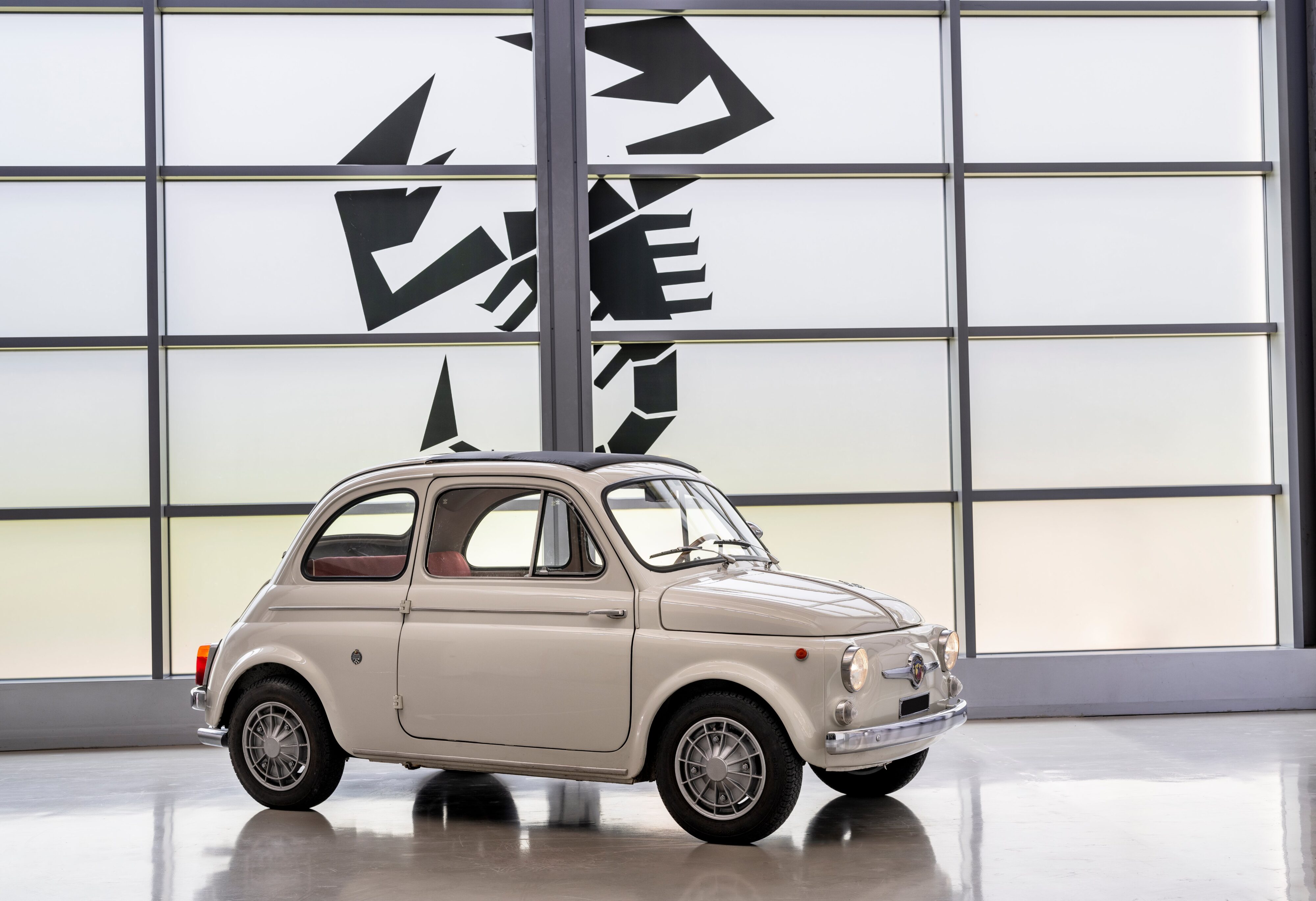 A photo of a 1963 Abarth 595 parked in front of a large Scorpion symbol, the emblem of the brand.