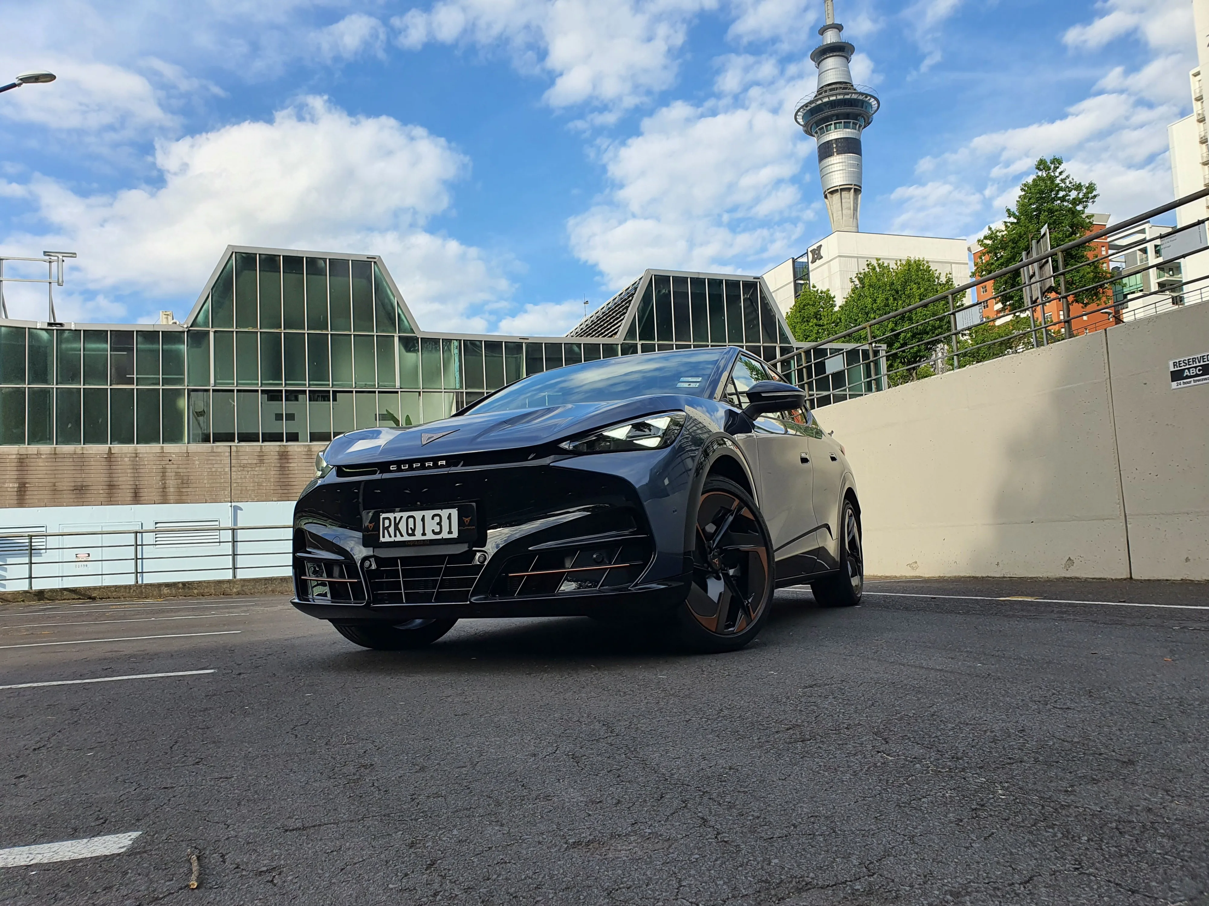 Front three quarters view of a 2025 Cupra Tavascan VZ in Tavascan Blue with the Auckland Sky Tower in the background.