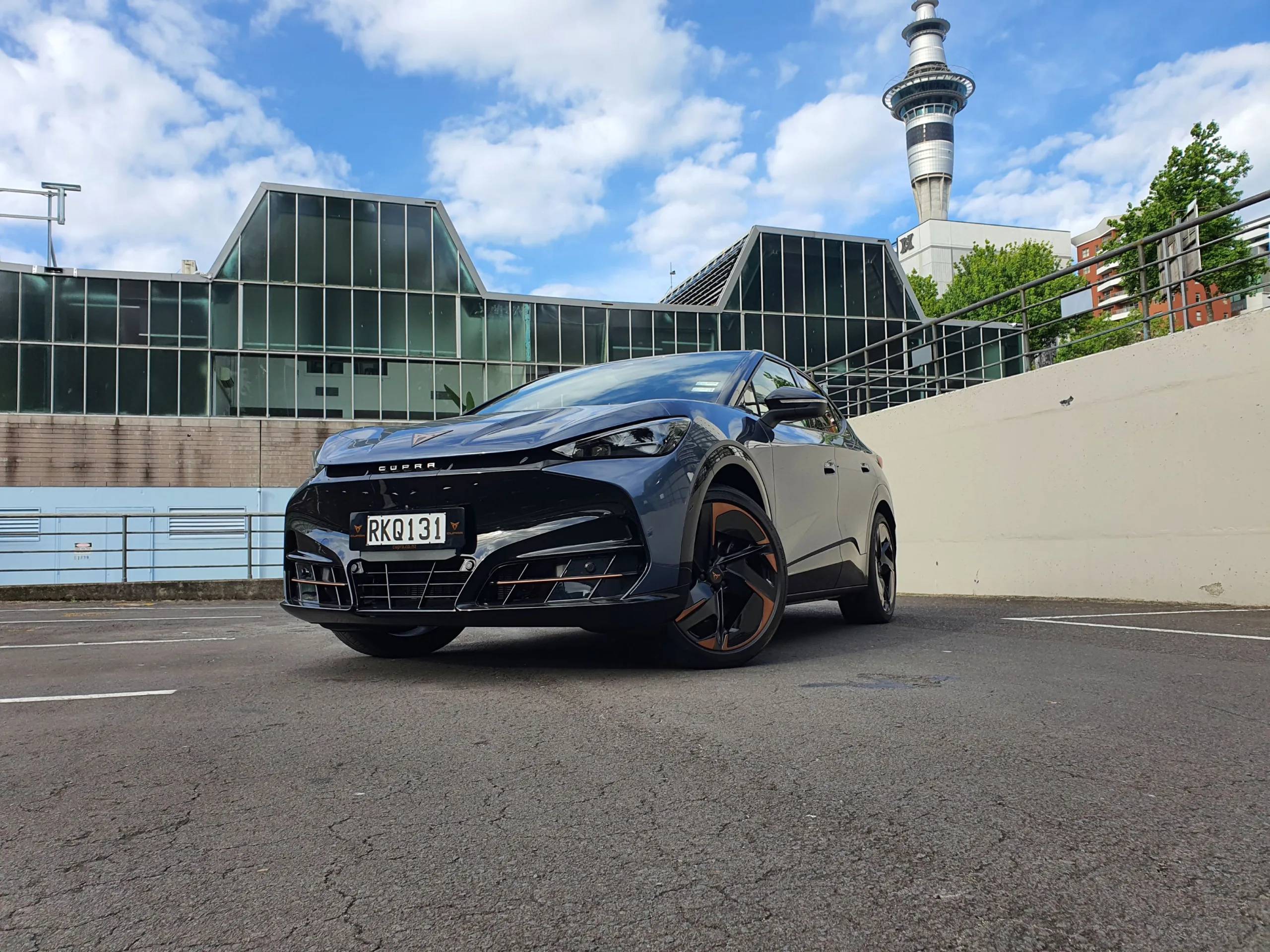 Front three quarters view of a 2025 Cupra Tavascan VZ in Tavascan Blue with the Auckland Sky Tower in the background.