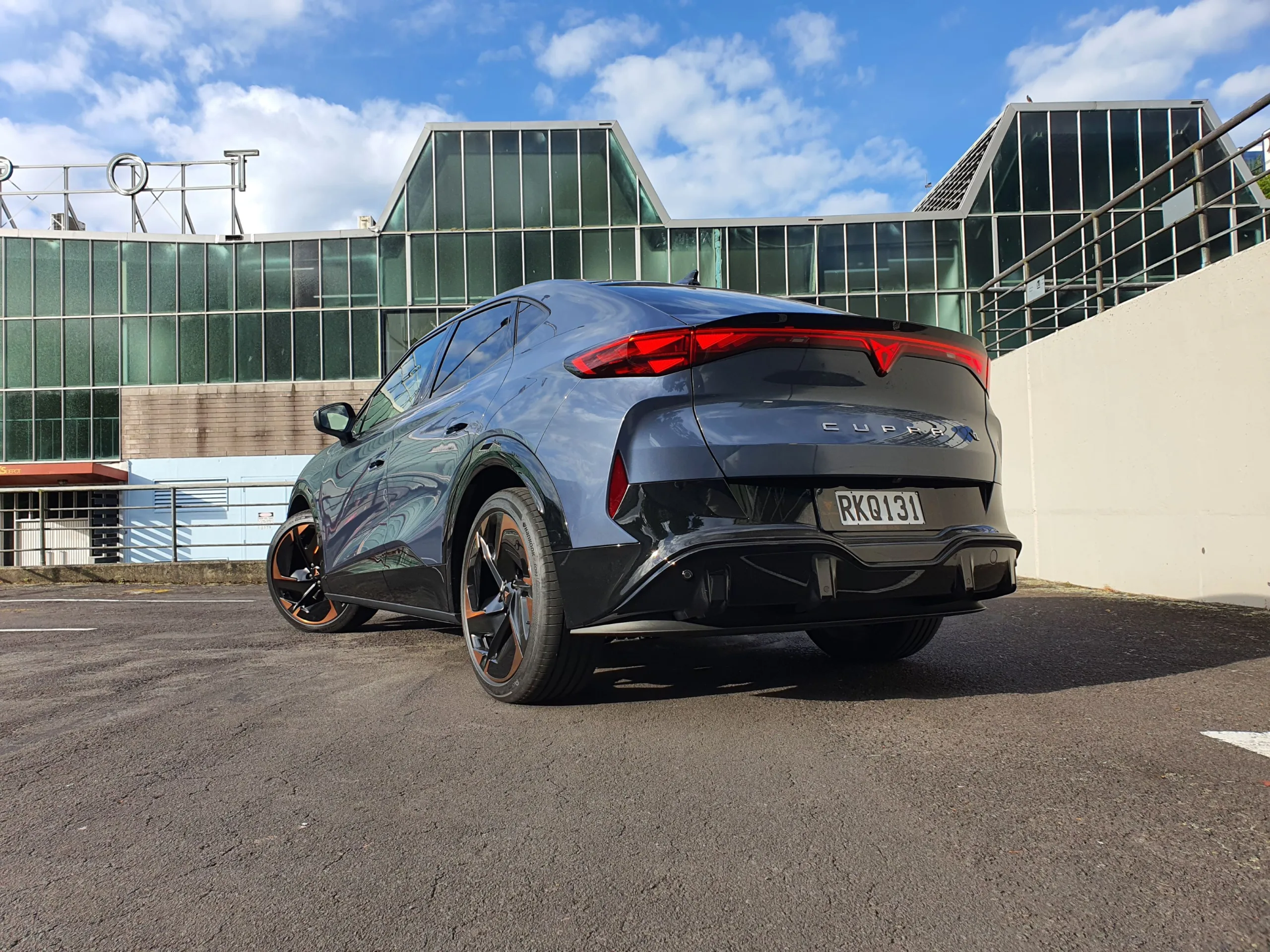 Rear three quarters view of a 2025 Cupra Tavascan VZ in Tavascan Blue with City Works Depot in the background.