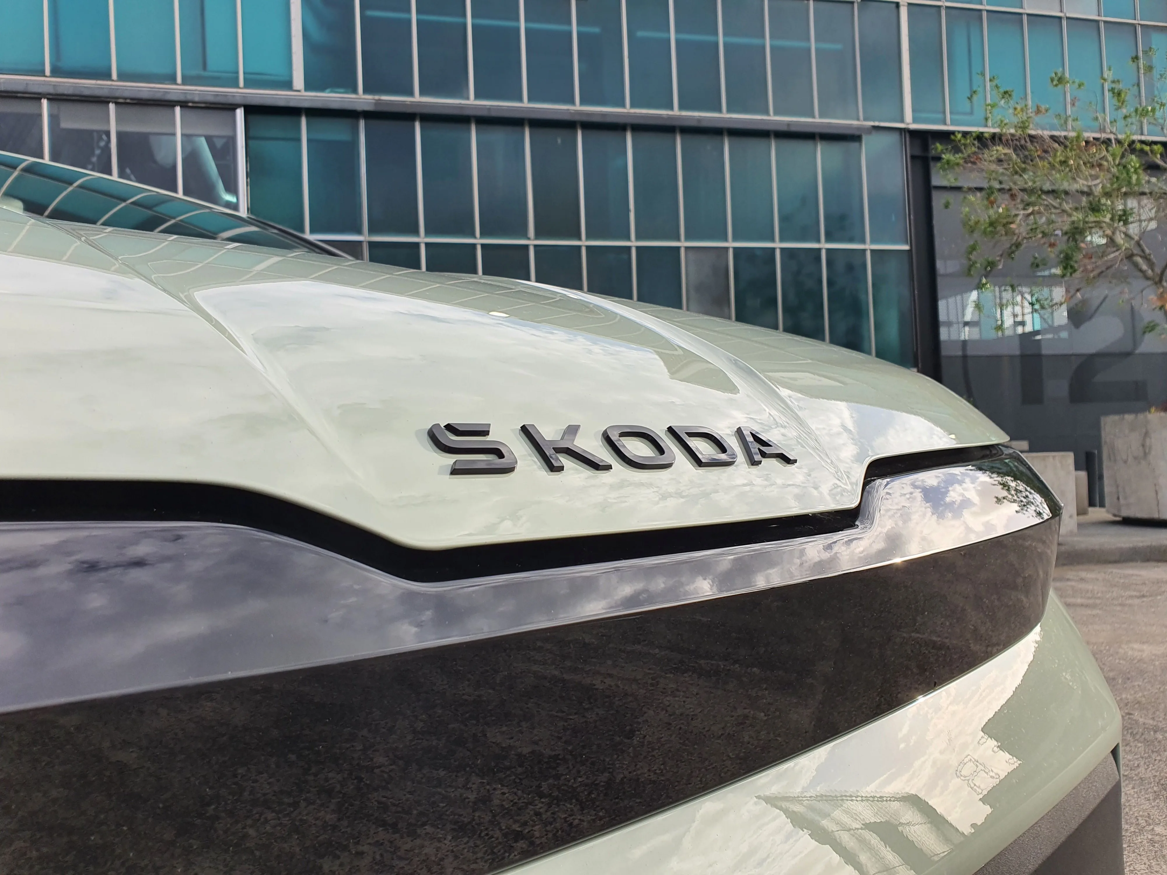 Close up view of the front badge on a 2026 Skoda Elroq Sportline in Timiano Green in front of an industrial building.