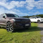 A photo of two Volvo EX90s near a beach just outside of Paihia, New Zealand. Car in the foreground is Mulberry Red, car in the background is Crystal White.