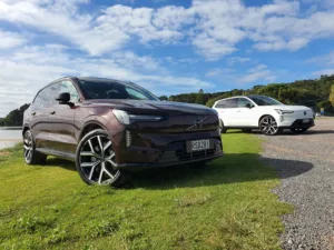 A photo of two Volvo EX90s near a beach just outside of Paihia, New Zealand. Car in the foreground is Mulberry Red, car in the background is Crystal White.