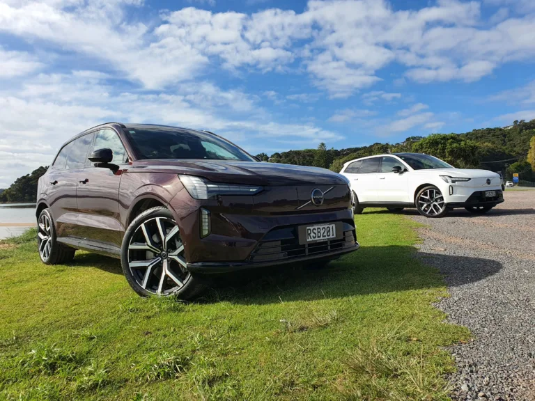 A photo of two Volvo EX90s near a beach just outside of Paihia, New Zealand. Car in the foreground is Mulberry Red, car in the background is Crystal White.