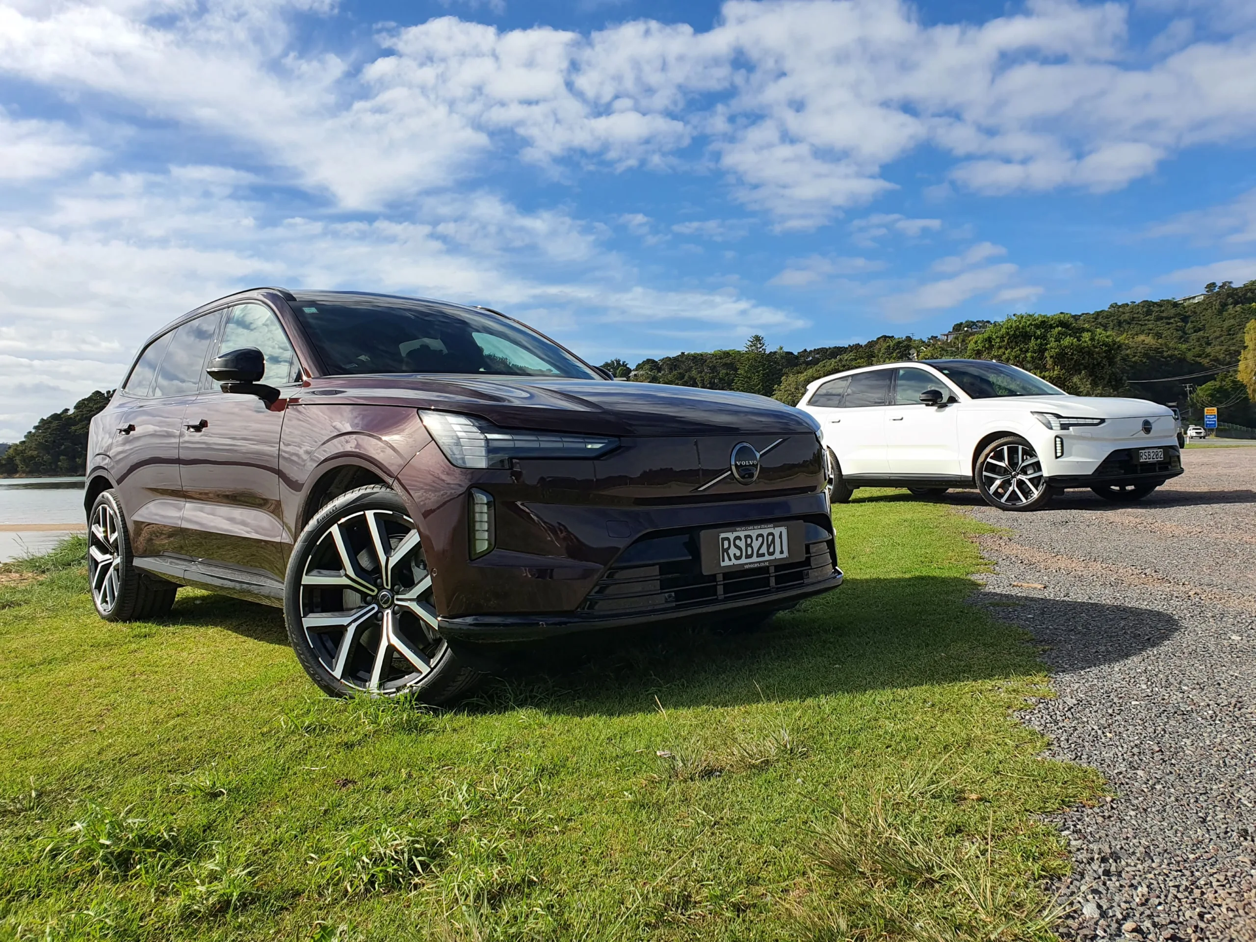 A photo of two Volvo EX90s near a beach just outside of Paihia, New Zealand. Car in the foreground is Mulberry Red, car in the background is Crystal White.