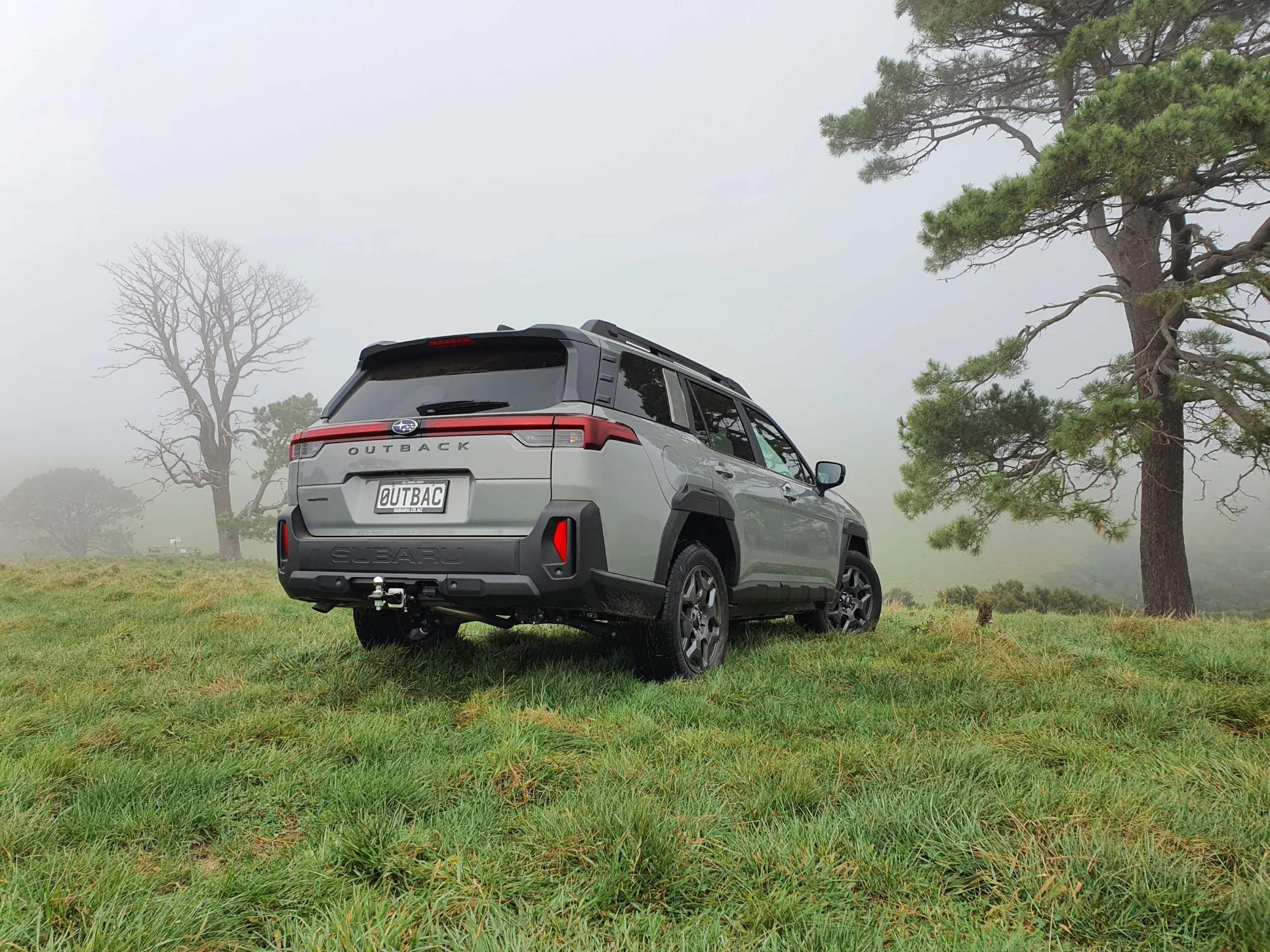Rear three quarters view of a 2026 Subaru Outback AWD in River Rock Pearl with a forest in the background.