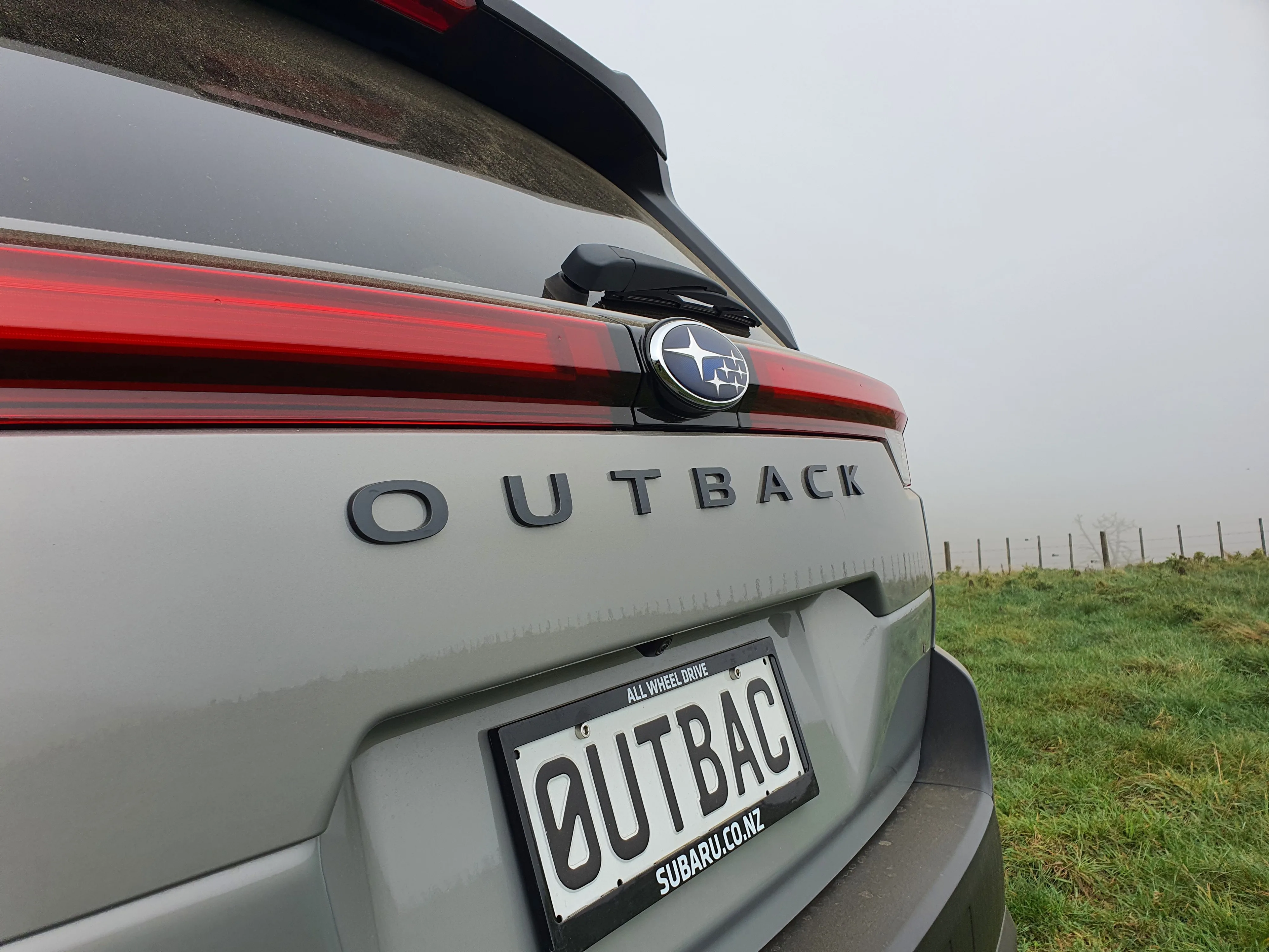 Close up of the Outback logo/lettering on the rear of a 2026 Subaru Outback AWD in River Rock Pearl with a forest in the background.
